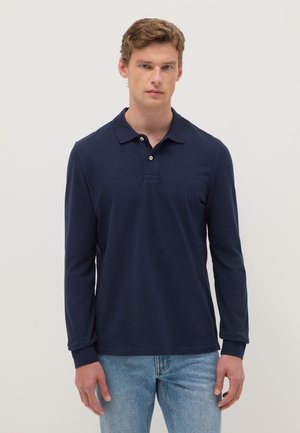 Hombre vestido con una camisa polo azul marino de manga larga y vaqueros azul claro, de pie frente a un fondo liso y claro.