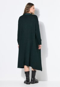 Lange, donkergroene sweater jurk van pluizig breimateriaal. Heeft een losse pasvorm met lange mouwen en een zoom die onder de knieën valt.