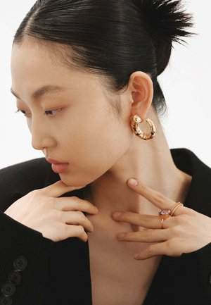 Pendientes de aro dorados con superficie texturizada y un acento redondo. La modelo lleva un anillo delicado con una piedra preciosa púrpura. Se ve un blazer negro.