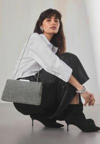 Vrouw hurkend, gekleed in een witte blouse, zwarte rok, zwarte laarzen met hak, zilveren armbanden, opvallende oorbellen en met een handtas met strass-steentjes.