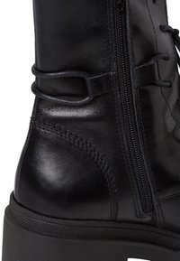 Gros plan d'un talon de bottine en cuir noir avec des coutures apparentes, une fermeture éclair latérale et des lacets décoratifs enroulés autour de la cheville.