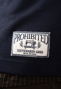 Ετικέτα ρούχου σε σκούρο ύφασμα με κείμενο "PROHIBITED," μια εικονογράφηση μιας vintage ραπτομηχανής, και "TEXTILWAREN GMBH Berlin, Germany."