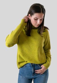 Junge vrouw met donker haar, gekleed in een groene trui en blauwe spijkerbroek, die met één hand haar haar aanraakt terwijl ze haar andere hand in haar zak heeft.