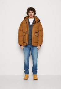 Brun pufferjakke med hætte og sorte detaljer, båret over en grå cardigan. Blå jeans og brune støvler fuldender outfittet.