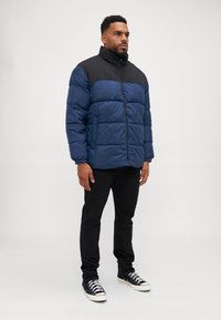 Doudoune bleue et noire avec une fermeture éclair, présentant un col haut, une texture matelassée et des poches latérales, associée à un pantalon noir et des baskets.
