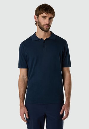 Uomo con barba che indossa una polo a maniche corte blu navy e pantaloni abbinati, in piedi davanti a uno sfondo chiaro e uniforme.