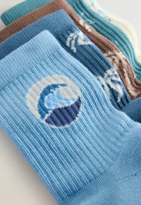 Pila de calcetines acanalados en azul, marrón y crema con diseños bordados de olas del océano y palmeras en la parte superior.