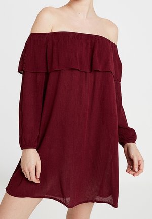 Kvinde iført en langærmet, off-shoulder, mørkerød kjole med flæsedetalje over brystet.