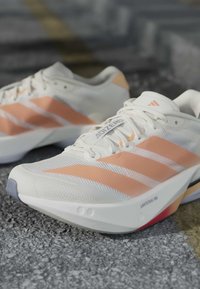 Λευκά και ροδάκινα παπούτσια Adidas για τρέξιμο με σόλα Lightstrike Pro πάνω σε υφή επιφάνειας.