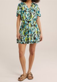 Kvinna i en kort klänning med bälte som har abstrakta bladmönster i blått, grönt och brunt, samt tanfärgade sandaler med två spännen.