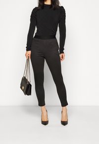 Svart långärmad topp med rynkade axeldetaljer, matchad med figurnära svarta rutiga byxor. Modellen håller en svart handväska med kedjestropp.