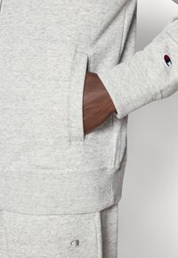 Ανοιχτό γκρι φούτερ με μπροστινή τσέπη, ριμπ μανσέτες και μικρή λεπτομέρεια λογότυπου. Μαλακό, υφασμάτινο με υφή και χαλαρή εφαρμογή.