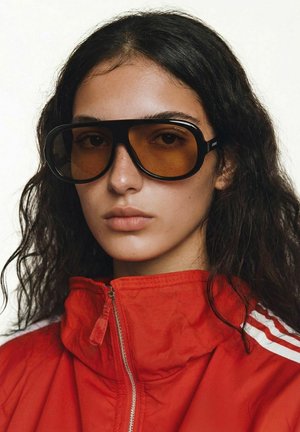 Jeune femme aux longs cheveux noirs portant de grandes lunettes noires à verres teintés orange et une veste rouge zippée avec des rayures blanches.