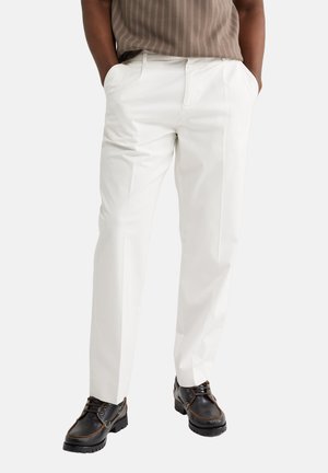 Fehér, testre szabott nadrágot, barna csíkos felsőt és fekete fűzős cipőt viselő férfi, kezeit a zsebében tartva áll.