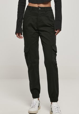 Personne portant un pantalon cargo noir avec des poignets élastiques et des baskets blanches, un haut noir à manches longues raccourci, debout devant un fond uni.