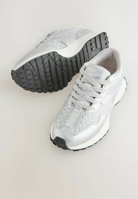 Srebrne sportowe sneakersy z akcentami z cyrkonii, z siateczkowym i syntetycznym materiałem, jasnoszarymi sznurówkami oraz teksturowaną czarną gumową podeszwą.