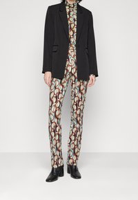 Zwarte blazer over een multikleurige jumpsuit met een geometrisch patroon in bruin, lichtblauw en crème. De outfit heeft een getailleerde broek en enkellaarsjes.