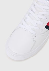 Witte leren sneaker met platte witte veters, ronde neus, gestructureerde rubberen zool en rode en marineblauwe strepen langs de zijkant. Klassiek ontwerp.