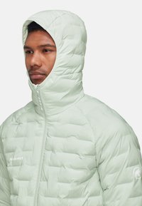 Helle minzgrüne Steppjacke mit hohem Kragen und integrierter Kapuze, mit gesteppte Abschnitte und einem Frontreißverschluss. Logo auf der Brust sichtbar.