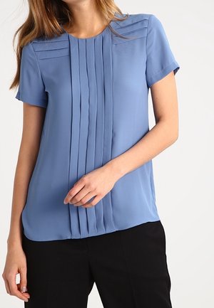 Kobieta ubrana w niebieską bluzkę z krótkim rękawem z pionowymi plisami z przodu, w połączeniu z czarnymi spodniami, na białym tle.