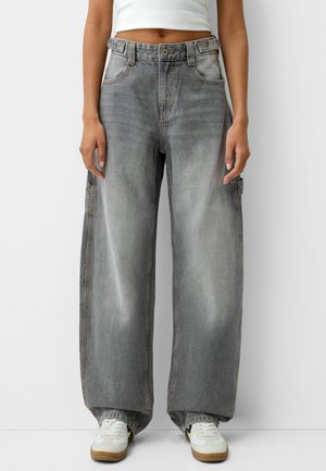 Persona con jeans anchos gris desgastado, top blanco corto y zapatillas blancas con suelas de goma, de pie contra un fondo blanco.