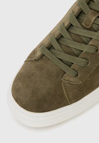 Olive green suede sneaker with round toe, flat white rubber sole, and matching laces. Features stitched accents along the sides.