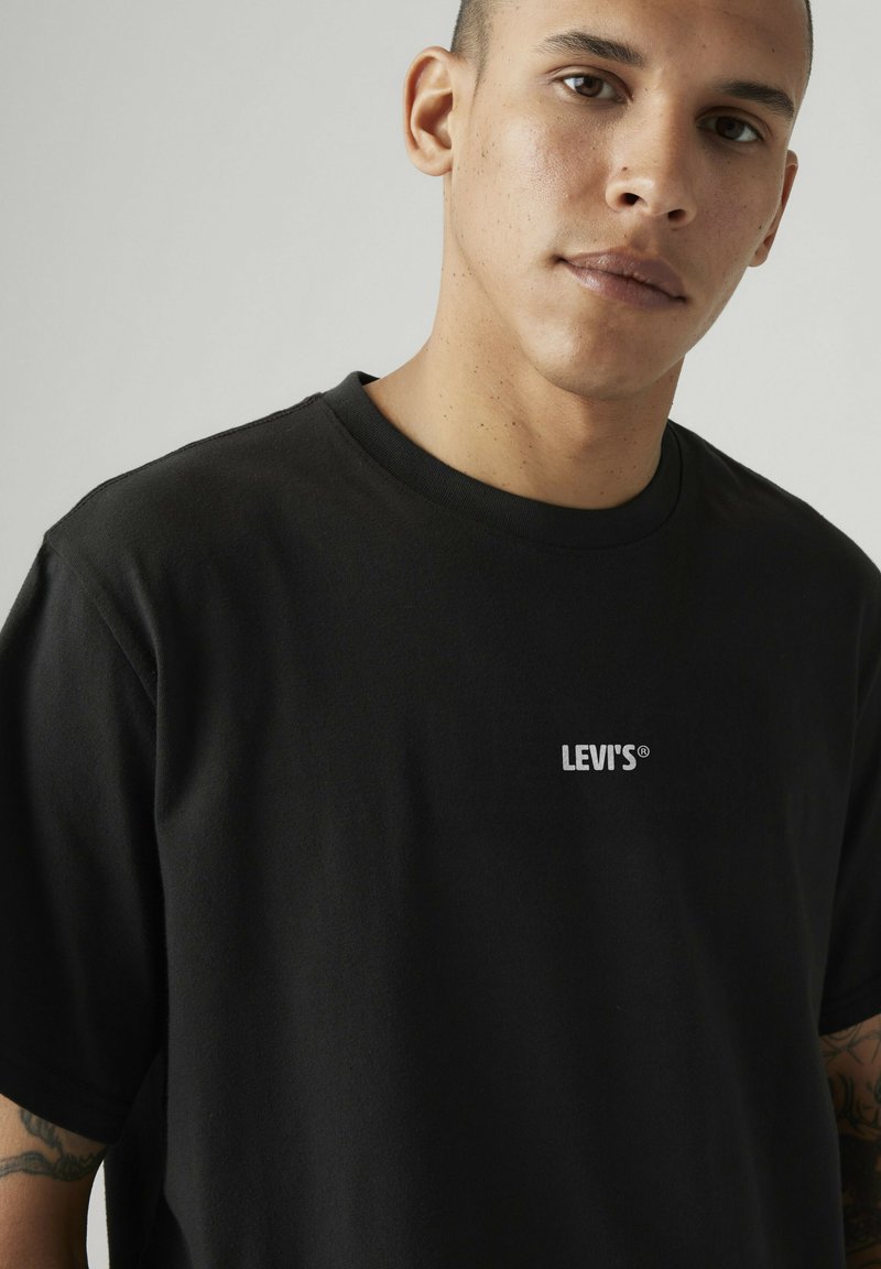 Joven con el pelo corto lleva una camiseta negra de Levi’s, mostrando tatuajes visibles en su brazo izquierdo, sobre un fondo gris liso.