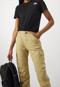 Svart kortärmad t-shirt med logotyp, tan cargobyxor med fickor, och en svart ryggsäck med remmar. Slät tygtextur.