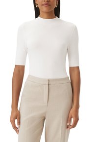 Haut blanc ajusté à manches courtes en tissu lisse et extensible avec un col montant. Associé à un pantalon beige sur mesure avec un détail de pli.