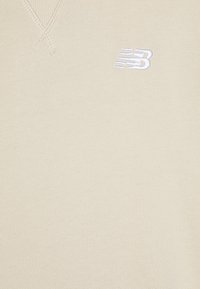 Világos bézs pulóver texturált kötött mintával, V-nyakkivágással és a bal mellen hímzett fehér "NB" logóval.