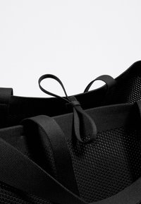 Zwarte mesh strandtas met textuurstof, voorzien van lusvormige straps en een gestructureerde vorm. Opvallend door zijn minimalistisch ontwerp en stevige constructie.