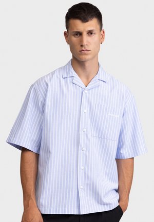 Hombre con camisa de manga corta azul claro a rayas verticales blancas y un bolsillo en el pecho, de pie contra un fondo liso.