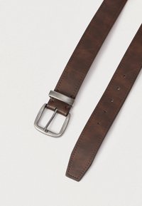 Καφέ δερμάτινη ζώνη με μεταλλική αγκράφα και πέντε τρύπες ρύθμισης σε ανοιχτό γκρι φόντο.