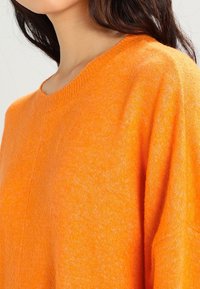 Pull en tricot orange avec un col rond, une coupe décontractée et un tissu légèrement texturé. Détails de couture visibles sur l'épaule.
