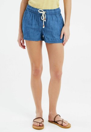 Shorts en denim bleu avec taille élastique, fermeture à cordon et poches latérales. Modèle portant des sandales brunes à lanières.