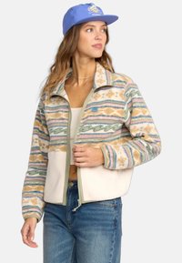 Chaqueta de forro polar con patrones geométricos multicolores, con una sección inferior en crema, cierre de cremallera y cuello alto. Acentuada con una etiqueta azul.