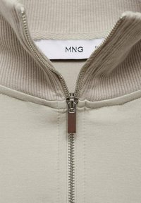 Giacca beige con zip e collare a costine, caratterizzata da una zip argentata e un'etichetta in tessuto con il logo "MNG". Tessuto morbido con un design minimale.