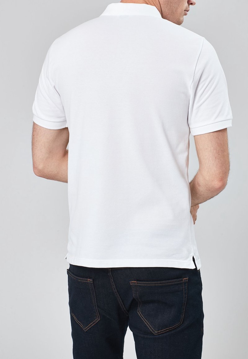 Homme portant un polo blanc simple à manches courtes et un jean bleu foncé, de dos contre un fond neutre.
