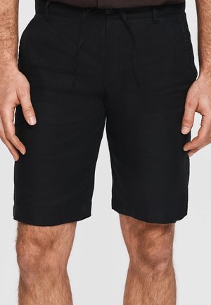 Czarne szorty do kolan noszone przez osobę stojącą z rękami swobodnie opuszczonymi wzdłuż ciała, pokazujące przednie kieszenie oraz szczegóły guzika.