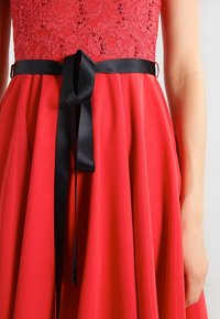 Robe rouge avec un corsage en dentelle ornée de sequins, une jupe en satin évasée et un ruban en satin noir noué à la taille.