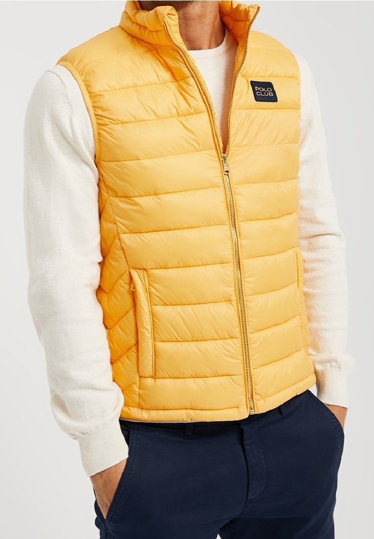 Gul vadderad väst med hög krage, horisontell sömnad, dragkedjestängning och en svart logotyp på bröstet. Bärs över en vit tröja.