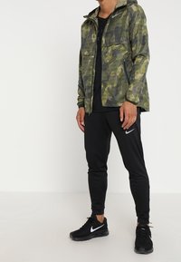 Man in camouflagejas, zwarte Nike joggers en zwarte Nike sneakers, staand tegen een effen achtergrond.