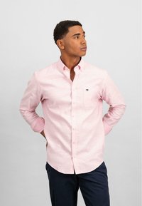 Camicia button-up rosa chiaro a maniche lunghe con taschino sul petto e piccolo logo scuro. Tessuto morbido e orlo curvo, abbinata a pantaloni scuri.