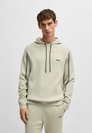 Felpa leggera color beige chiaro con dettagli e logo neri, dotata di cappuccio con coulisse e tessuto testurizzato, indossata con pantaloni coordinati.