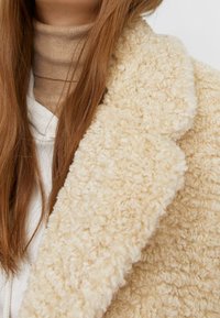 Lange braune Haare über einem beigen Rollkragenpullover und einem cremefarbenen, strukturierten Wollmantel mit Kragen und Revers.