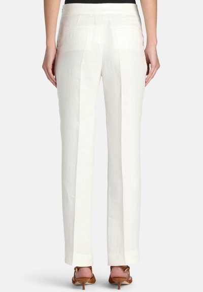 Nő fehér, magas derekú, hátsó zsebes, szabott nadrágban és barna, magas sarkú szandálban, egy egyszerű világos háttér előtt áll.