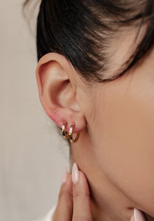 Orecchini a cerchio dorati con una finitura lucida, caratterizzati da un design a doppio cerchio. Vista ravvicinata dell'orecchio, che mostra la pelle liscia e la texture dei capelli naturali.