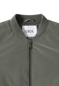 Oliivivihreä pommikurki, jossa on ribbetty kaulus ja edessä vetoketju, näkyy valkoinen etiketti, jossa lukee "CECIL" brändin nimi kaulusessa.