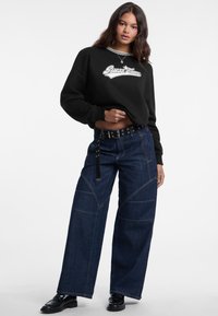 Donna che indossa una felpa nera con il logo "Guess Jeans", jeans a gamba larga blu scuro con cintura e mocassini neri, in piedi contro uno sfondo bianco.