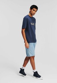 T-shirt navy avec le logo "KARL", associé à un short à motifs bleu clair. Le modèle porte des baskets montantes noires avec des semelles blanches et des chaussettes à logo.